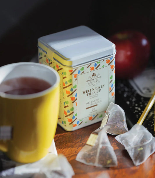 Wellness in the Cup tea tin with herbal tea sachets, yellow mug, and red apple on a wooden table